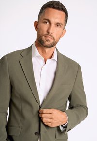 Olijfgroen op maat gemaakte blazer met een notch lapel, enkele knoop en zakken. gedragen over een witte blouse met een gespreide kraag en minimale knopen.
