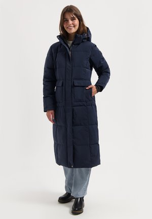 Frau lächelt, trägt einen langen, marineblauen Daunenmantel mit Kapuze, helle blaue Jeans und schwarze Stiefeletten, steht vor einem schlichten weißen Hintergrund.