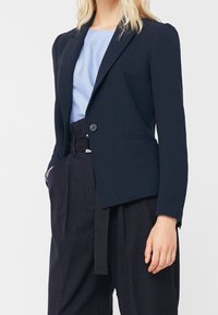 Blazer bleu marine en tissu lisse, avec un bouton unique, un col à revers, et une coupe ajustée, associé à un haut bleu clair et un pantalon rayé foncé.