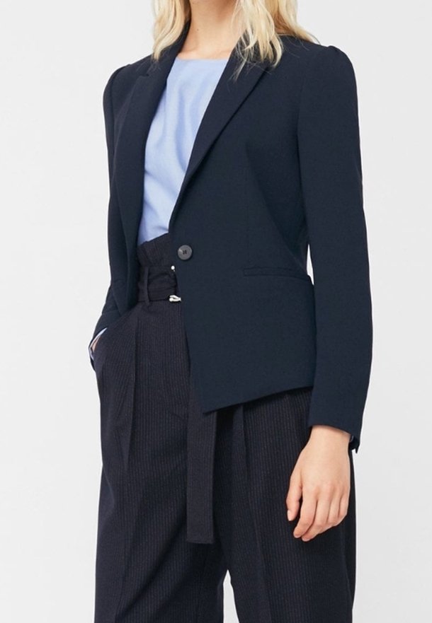 Blazer bleu marine en tissu lisse, avec un bouton unique, un col à revers, et une coupe ajustée, associé à un haut bleu clair et un pantalon rayé foncé.