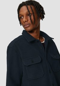 Dunkelblaues Jacket aus strukturiertem Stoff, mit Hemdkragen, zwei vorderen Taschen mit Klappen und schwarzen Knöpfen.