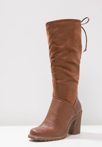 Botte haute marron jusqu'au genou avec une tige en daim souple, un bout en cuir lisse, une forme de bout arrondi et un talon haut épais. Détail de semelle texturée.