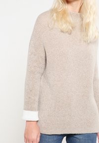 Beige Strickpullover mit lässiger Passform, ausgestattet mit einer strukturierten Oberfläche und weißen Ärmelakzenten. Das Design hat einen runden Ausschnitt und lange Ärmel.
