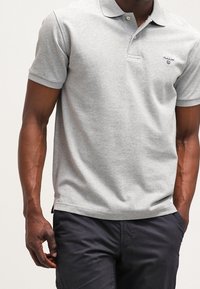 Grå polo-shirt i bomull med klassisk krage, korta ärmar och en liten broderad logotyp på vänster bröst. Bärs med mörka byxor.