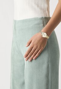 Light green corduroy pants with vertical ribbing, paired with a silver-tone watch featuring a white face and link bracelet. Hand resting on hip.