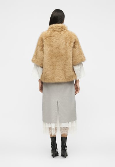 Femme portant une veste courte beige duveteuse sur un haut blanc à manches longues, jupe grise avec fente dans le dos, ourlet en dentelle et bottines noires, de dos.