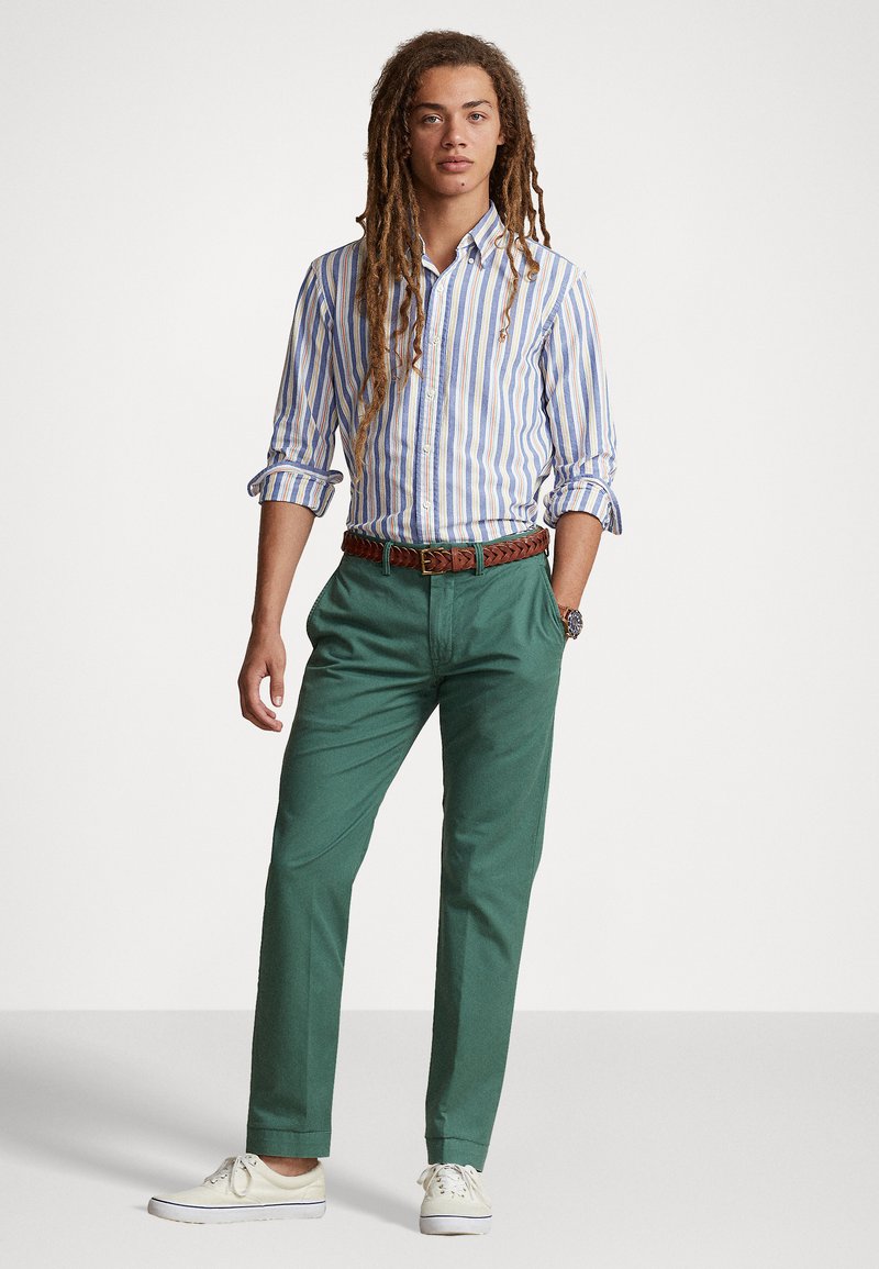 Gestreiftes Hemd in Blau und Weiß mit orangen Akzenten, kombiniert mit grünen Chinos und einem braunen geflochtenen Gürtel. Weiße Schuhe runden den Look ab.