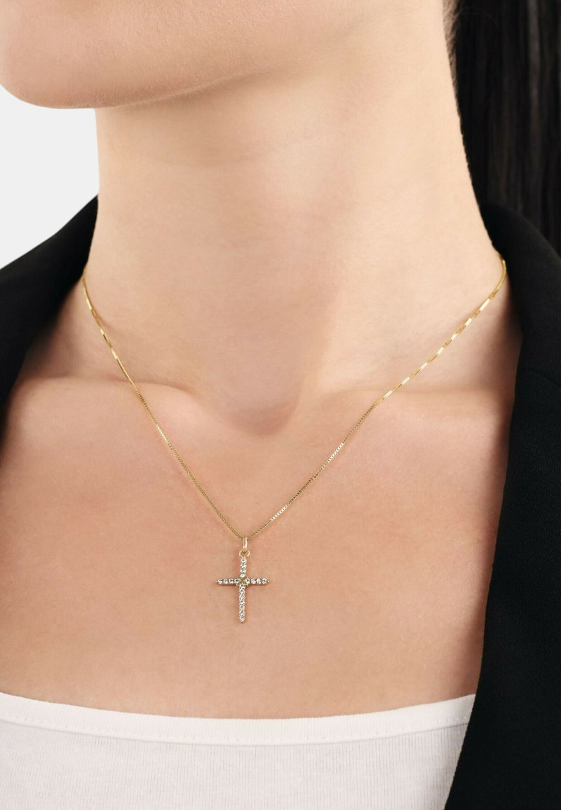 Collana d'oro con una delicata catena e un pendente a croce adornato con gemme trasparenti, poggiata appena sopra una clavicola nuda.
