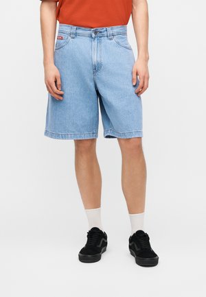 Mannelijk model draagt lichtblauwe spijkershorts, witte sportsokken, zwarte sneakers en een oranje shirt, staand tegen een effen achtergrond.