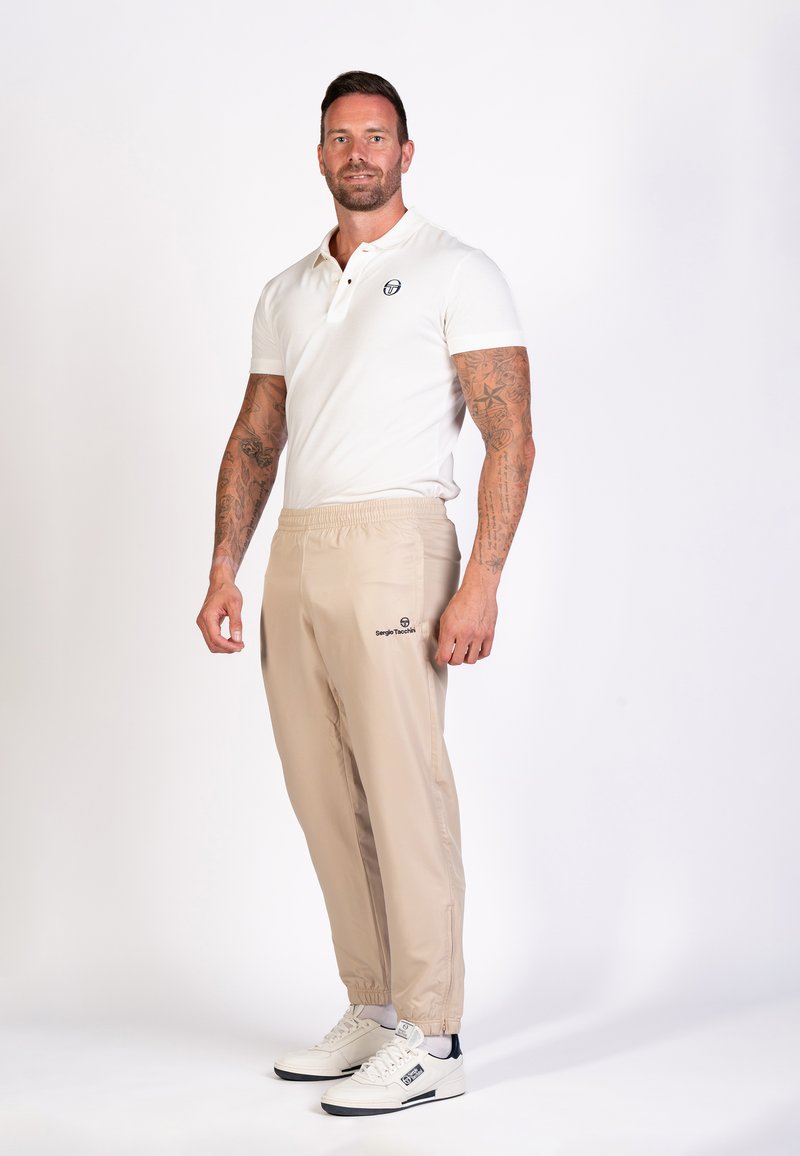 Modelo vistiendo una camiseta polo blanca, pantalones de cinturilla elástica beige y zapatillas blancas con detalles oscuros. Los pantalones tienen un logo en el lado.