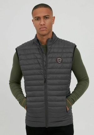 Grau gesteppte Weste mit vertikalem Quilt-Design, hohem Kragen und durchgehendem Reißverschluss. Mit einem runden Logopatch auf der linken Brust.