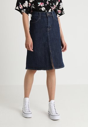 Kvinde iført en mørkeblå denimnederdel til knæet med en slids foran, hvide høje sneakers og en sort blomstret bluse.