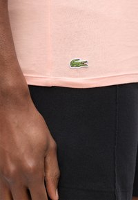 Χέρι με σκουρόχρωμο δέρμα που ακουμπά δίπλα σε ένα ανοιχτό ροζ πουκάμισο με πράσινο λογότυπο κροκόδειλου κοντά σε μαύρο παντελόνι.