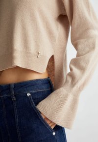 Beige lyhyempi neule, jossa on leveät hihat, tummansinisien korkeavyötäröisten farkkujen päällä, käsi etutaskussa, paljastaen vatsan.