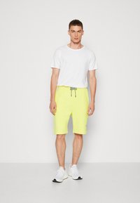 Weißes Baumwoll-T-Shirt kombiniert mit hellgelben Shorts, die über einen Zugverschluss und graue Akzente verfügen. Das Modell trägt weiße Sportschuhe.