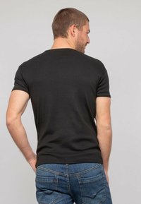 T-shirt noir à manches courtes en coton, avec un col rond. Le modèle porte un jean en denim bleu. L'arrière-plan est gris clair.