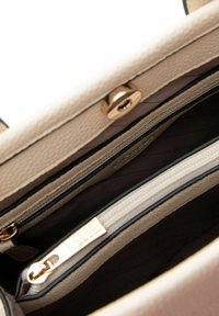 Interno di borsa beige in tessuto con hardware dorato, dotato di scomparti con zip e un'etichetta di marca. Fodera liscia completa il design.