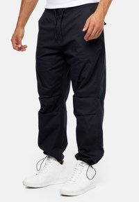 Sorte, afslappede cargo-bukser i en løs pasform med elastisk talje og snoretræk, der har flere lommer og lynlåsdetaljer. Bæres med hvide sneaks.