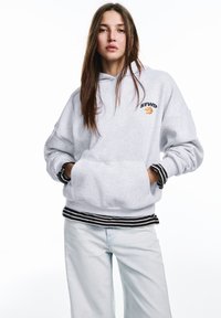 Hanorac gri cu glugă și buzunar frontal, având logo și grafică. Accentuate cu manșete și tiv cu dungi alb-negru. Purtați cu blugi albastru deschis.