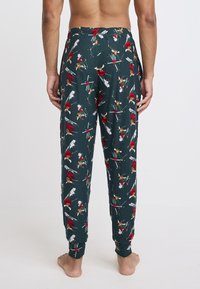 Groene pyjamabroek van zacht materiaal, met een speels ontwerp van rode en bruine cartoonfiguren die skiën op een witte achtergrond.