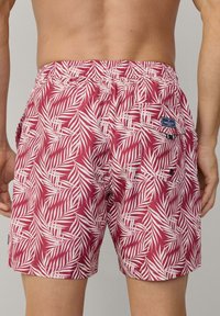 Pantaloni da bagno rossi con motivo a foglie di palma bianche, vita elastica e una tasca posteriore con patch logo. Tessuto morbido e leggero.