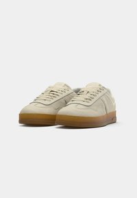 Paire de baskets en daim beige avec semelles en caoutchouc marron clair et lacets beiges sur fond gris clair uni.