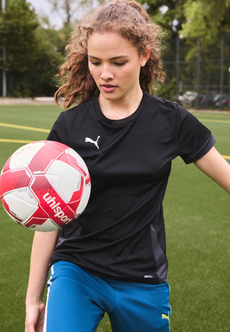 Black athletic shirt with short sleeves and silver logo, paired with blue athletic pants. The player balances a red and white soccer ball.