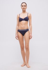 Γυναίκα που στέκεται ξυπόλητη μπροστά σε λευκό φόντο, φορά μπλε σκούρο σουτιέν και αντίστοιχο εσώρουχο Polo Ralph Lauren.