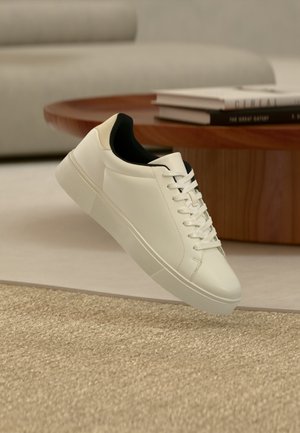 Baskets basses blanches avec doublure intérieure noire, lacets blancs et semelle texturée flottant au-dessus d'un tapis beige dans un salon moderne.