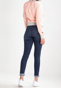 Žena v narolovaných tmavě modrých skinny džínách, světle růžové bomber bundě a bílých teniskách, stojící zády k bílé pozadí.