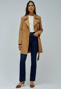 Abrigo marrón texturizado de doble botonadura con botones dorados, llevado sobre una blusa blanca y jeans azul oscuro, combinado con zapatos de tacón puntiagudos marrones.