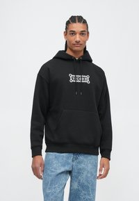 Haut à capuche noir avec une poche avant et un graphique blanc de "Volcom" à l'intérieur d'une bordure ornée, porté avec un jean texturé bleu clair.