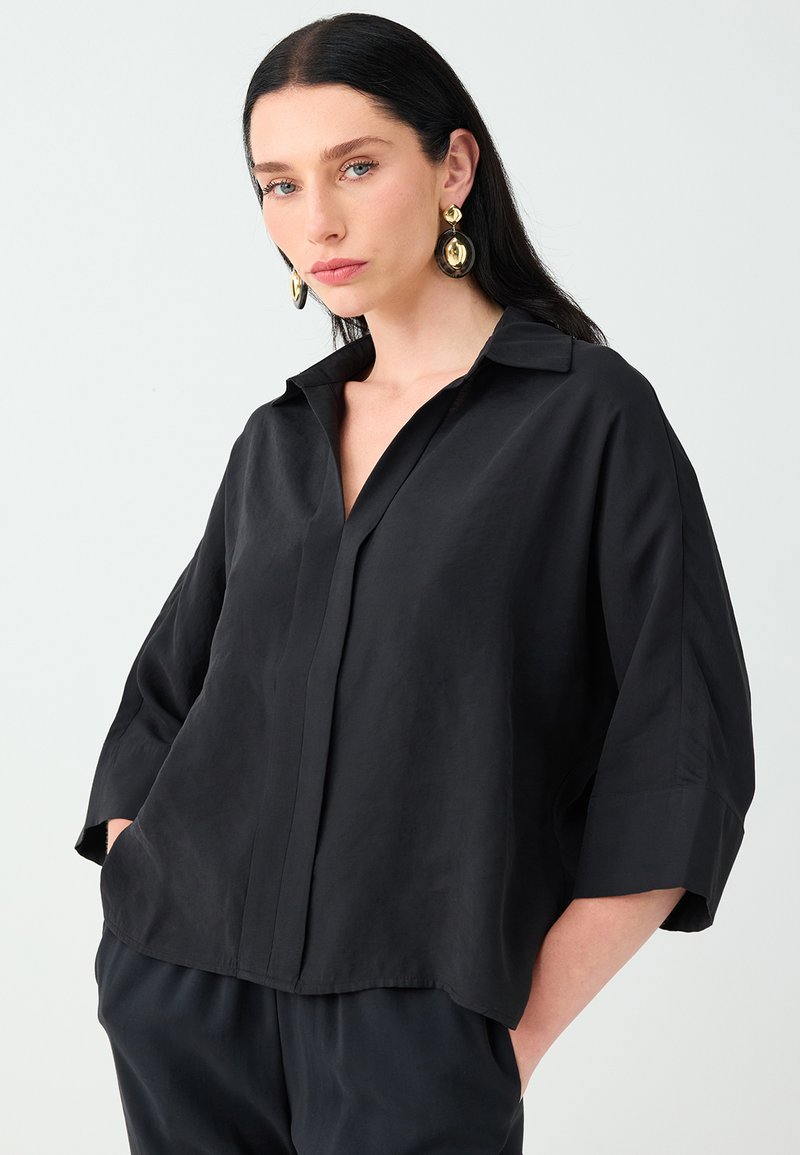 Femme aux longs cheveux foncés portant des boucles d’oreilles dorées et une chemise noire ample à col, les mains dans les poches, sur un fond clair uni.