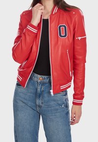 Kobieta w czerwonej skórzanej kurtce w stylu varsity z białymi paskami i literą "O", w połączeniu z czarnym topem i niebieskimi dżinsami.