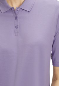 Lila Polo-Shirt mit strukturierter Oberfläche, drei Knöpfen am Kragen und kurzen Ärmeln. Der Stoff wirkt leicht und atmungsaktiv.