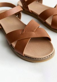 Bruna sandaler i konstläder med korsande remmar, en slät beige fotbädd och en sydd gummisula. Enkelt, öppet design.