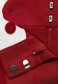 Red fabric with a textured finish; features a red button, two metallic clips, and a white section. Visible stitching and zipper details.