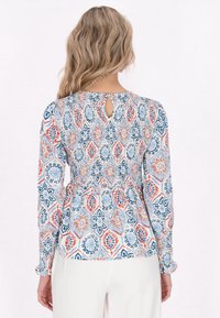 Bluse med multikolor mønster har lange ærmer, smocket talje og en nøglehuldetalje bagpå. Stoffet har en blød tekstur.