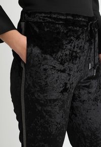 Svarta sammet-joggers med en mjuk textur, sidofickor och dragsko-stängning. En tunn grå rand löper längs sömmen på sidan.