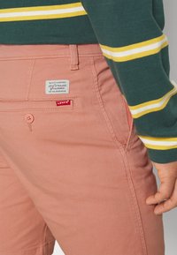 Shorts en coton rose avec une poche latérale, une étiquette de marque et un détail de bouton. Un haut vert à manches longues avec des rayures jaunes est partiellement visible.