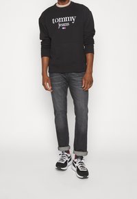 Uomo che indossa una felpa nera Tommy Jeans, jeans scuri arrotolati, scarpe sportive nere e bianche, in piedi contro uno sfondo chiaro e semplice.