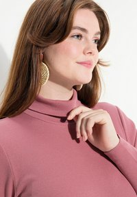 Rosa-pinkes, geripptes Shirt mit Mock-Turtleneck und langen Ärmeln. Getragen mit runden, gewebten beigefarbenen Ohrringen. Das Model berührt sanft den Kragen mit einer Hand.