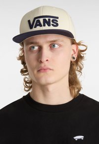 Kremowa czapka VANS z ciemnoniebieskim daszkiem, z dużym haftowanym logo w kolorze granatowym. Noszona z czarną koszulą z okrągłym dekoltem, na której znajduje się mała grafika deskorolki.