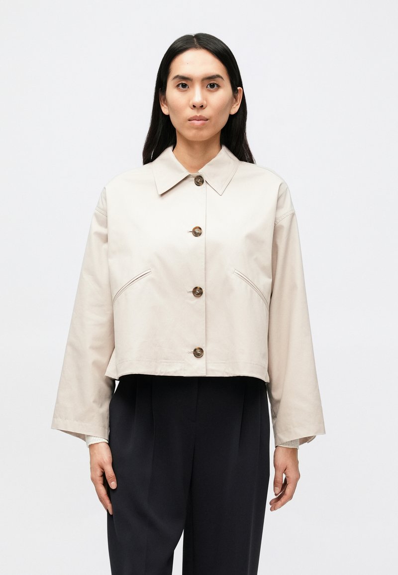 Κοντό μπεζ σακάκι από απαλό ύφασμα, με κλασικό γιακά, κλείσιμο με πέντε κουμπιά και δύο διαγώνιες μπροστινές τσέπες.