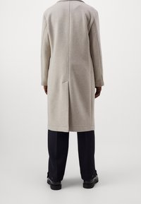 Manteau en laine gris clair, coupe droite, manches longues et détail de couture au dos, associé à un pantalon noir et des chaussures.