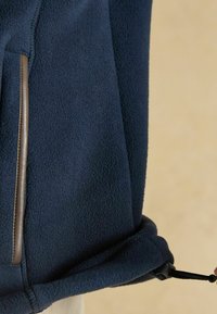 Detailný záber na námornícky fleecový kabát s vreckom s hnedým lemom a nastaviteľným šnúrkovým pásom na spodnej strane, ktorý je ťahaný rukou.