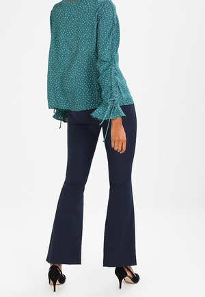 Taufelblaue Bluse mit Punkten, langen Ärmeln und Bindedetails, kombiniert mit schmalen, navyfarbenen Hosen und schwarzen High Heels.