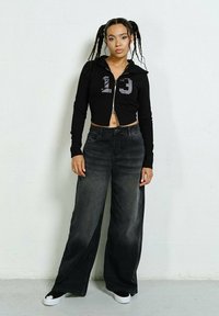 Czarna kurtka z suwakiem, krótka, z graficzną liczbą 13, w połączeniu z szerokimi szarymi dżinsami. Modelka nosi także czarne buty wsuwane.