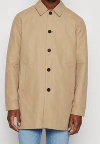 Beige jas met een sluiting aan de voorkant met knopen, een puntige kraag en twee zijzakken. Gemaakt van gladde stof met een iets losse pasvorm.