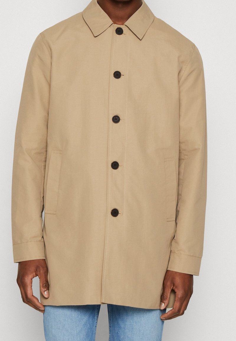 Beige jas met een sluiting aan de voorkant met knopen, een puntige kraag en twee zijzakken. Gemaakt van gladde stof met een iets losse pasvorm.